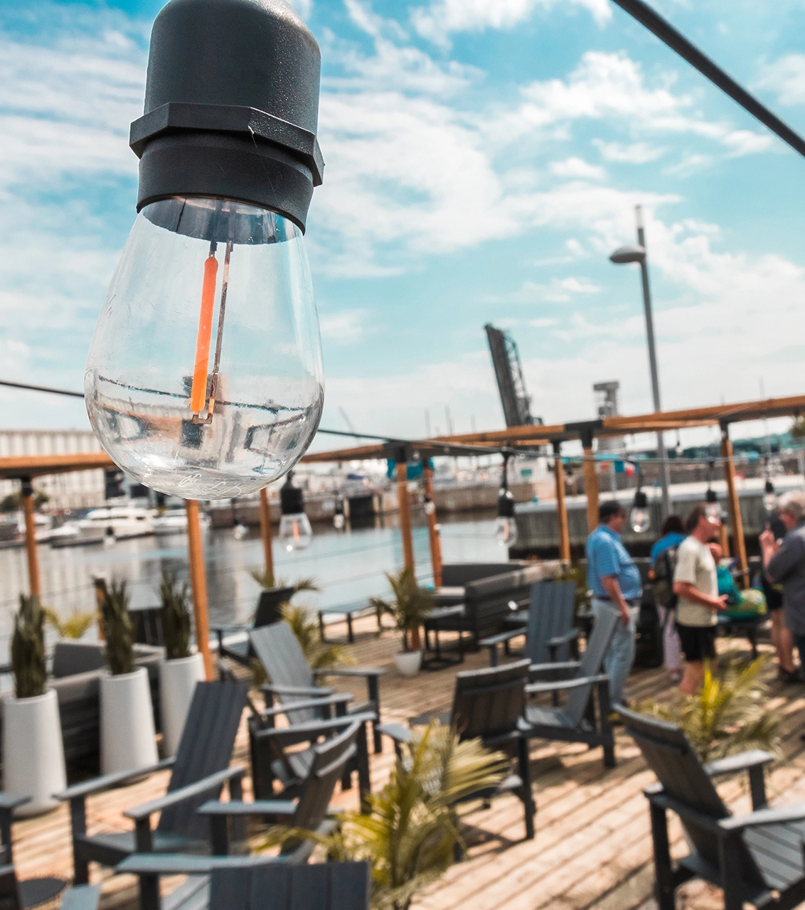 Close-up of a hanging light bulb with a waterfront lounge area and people gathered at the Oasis du Port de Québec.
